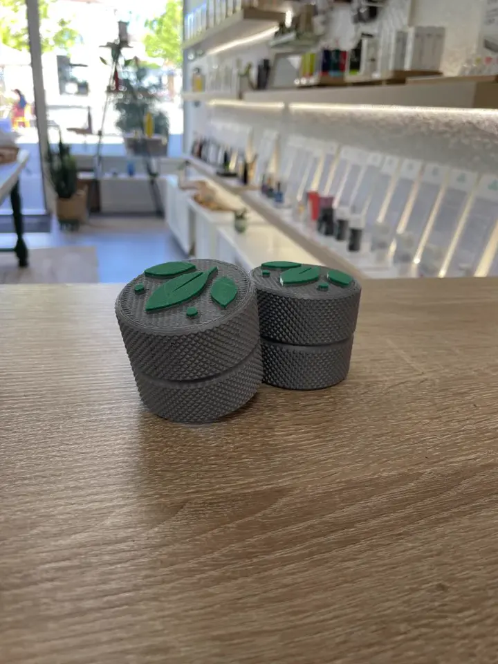 Petites boîtes rondes moletées grises, imprimées en 3D, au couvercle orné d’un logo vert, posées sur le comptoir d’une boutique