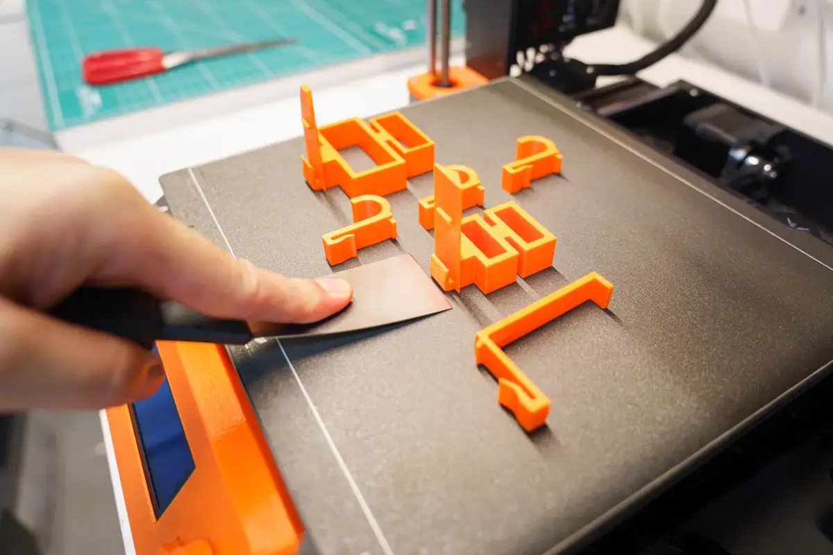 Main qui décolle des pièces orange du plateau d’une imprimante 3D avec une spatule